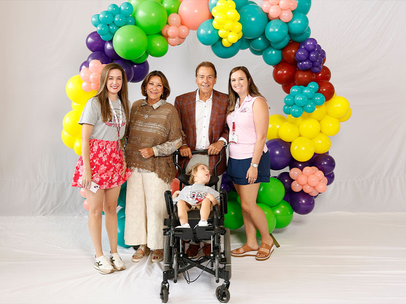Group photo of four adults and a child under colorful balloon arch, child seated in front.