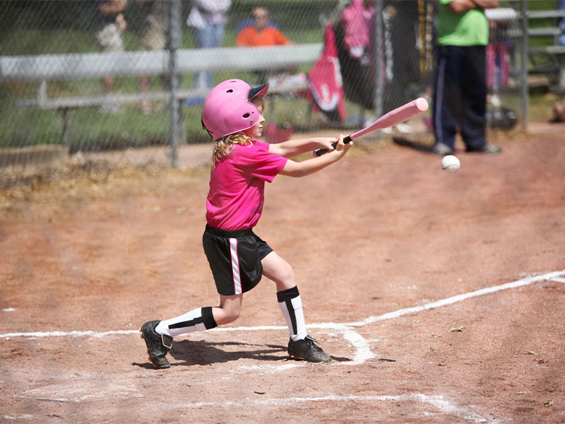 A young baseball player in a pink helmet swings at a pitch on a dusty field during a little league game.