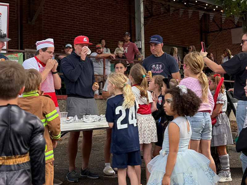Kids in costumes watch adults in a hot dog eating contest at an outdoor fair, with a table of foil-wrapped food.