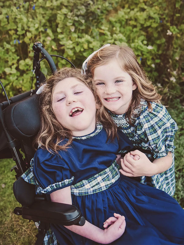 Two girls in blue dresses, one seated, smiling and hugging outdoors among greenery.