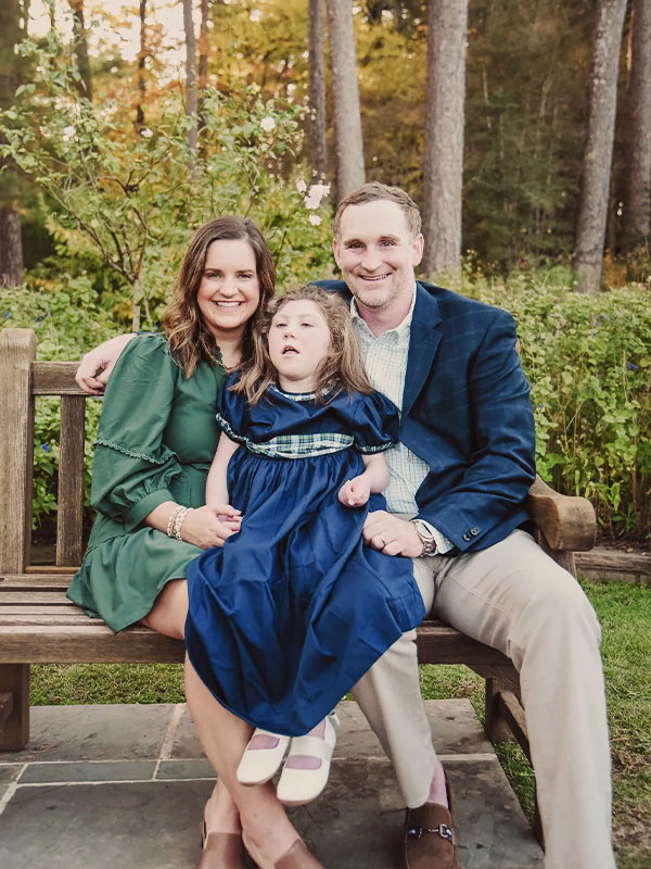 Family of three on a wooden bench in a garden; woman in green dress, girl in blue dress, man in blazer, trees behind.