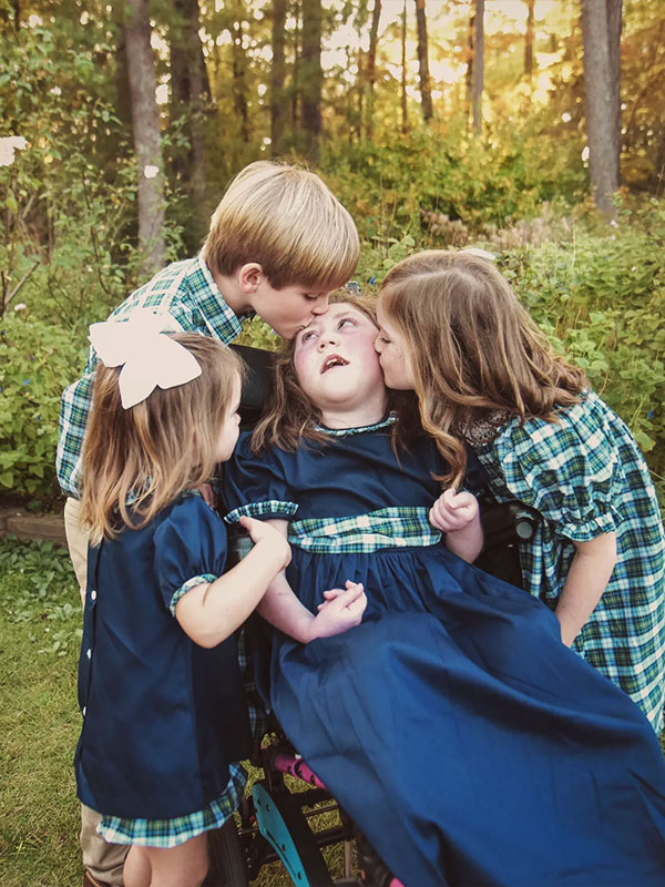 Three siblings hug and kiss a child in a blue dress outdoors at sunset, all wearing matching plaid outfits