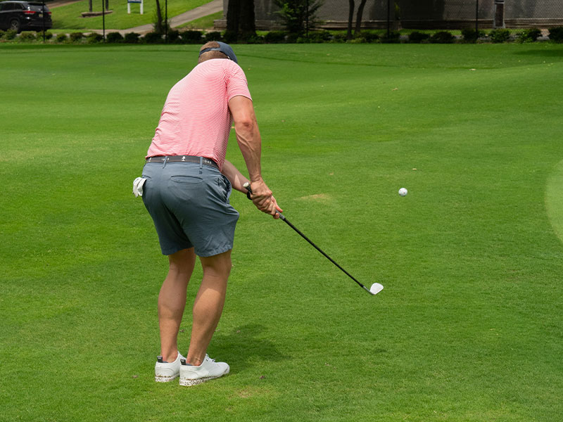 Golfer chipping a ball on a green fairway, wearing a pink striped shirt, blue shorts, white sneakers, and a cap.