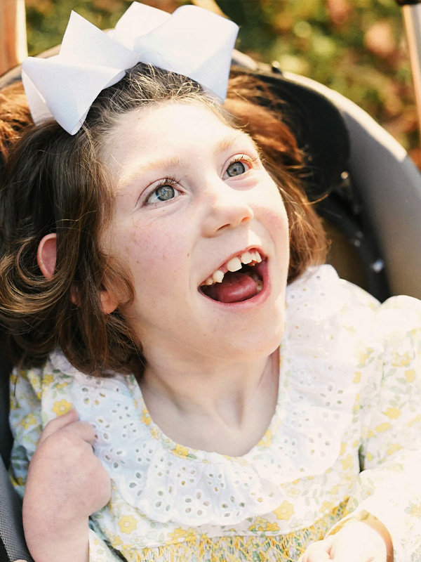Smiling child with white bow and floral dress with eyelet lace collar, looking up while seated outdoors