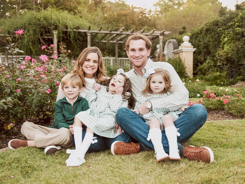 A family of five sits on grass in a rose garden, parents with three kids in green outfits, smiling outdoors.