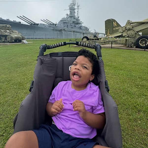 A child in a stroller at a military museum with a battleship, tanks, and artillery displayed on a grassy field.