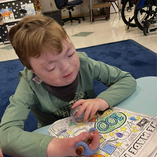 A child coloring a 100th day of school worksheet with crayons at a classroom table, blue carpet in background