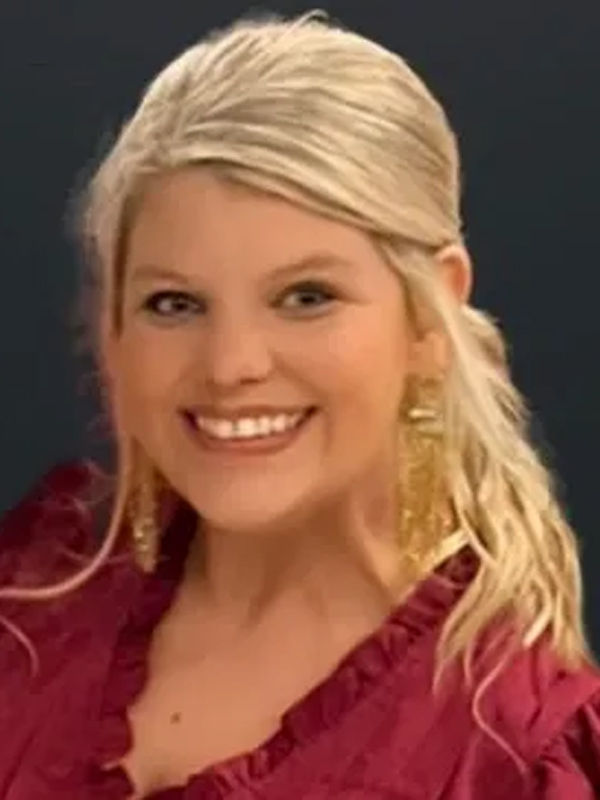 Smiling woman with blonde hair wearing maroon top and gold earrings, portrait on dark background
