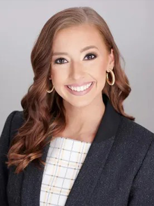 Professional headshot of a smiling woman with wavy brown hair, hoop earrings, and a dark blazer on a gray background