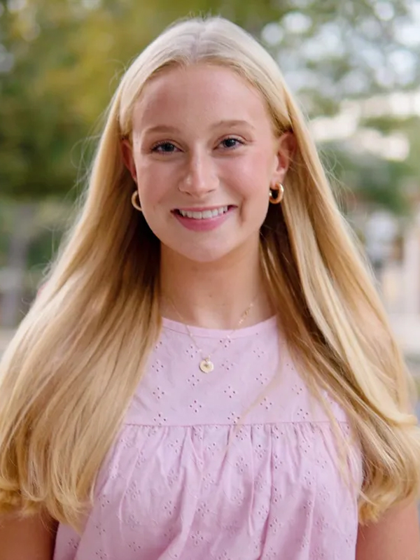 Smiling woman with long blonde hair in a pink blouse, outdoor portrait with soft background bokeh.