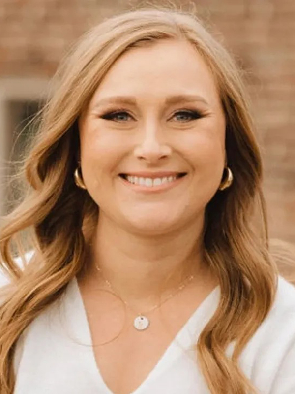 Smiling woman with long wavy hair wearing gold hoop earrings and pendant necklace in front of a brick wall