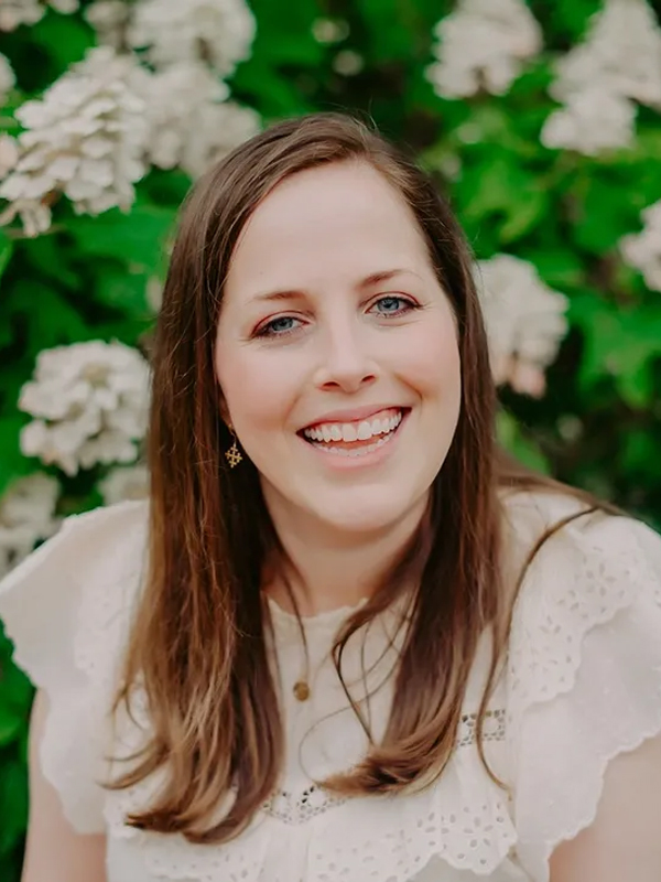 Smiling woman with long brown hair in white eyelet blouse, portrait by blooming hydrangeas.