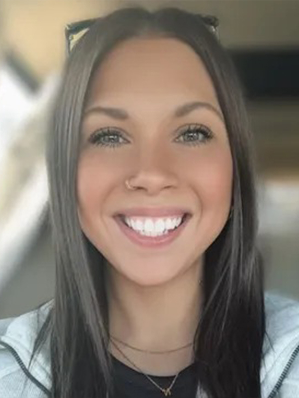 A woman with long dark hair and a nose ring smiling brightly, wearing layered necklaces and a light hoodie indoors.