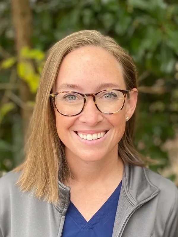 Close-up portrait of a smiling woman with glasses in blue scrubs and gray jacket outdoors