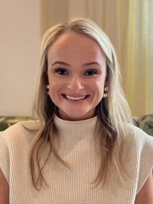 Smiling woman with long blonde hair and pearl earrings indoors, wearing a sleeveless ribbed turtleneck top.