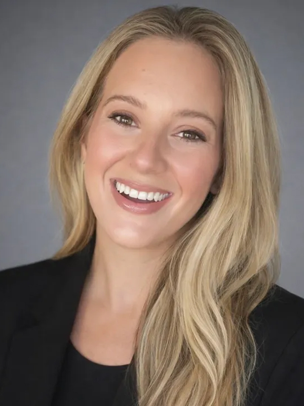 Smiling professional woman with long blonde hair in black blazer, studio headshot on gray background.