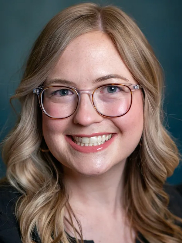 Smiling woman with blond wavy hair and glasses in professional headshot on teal background