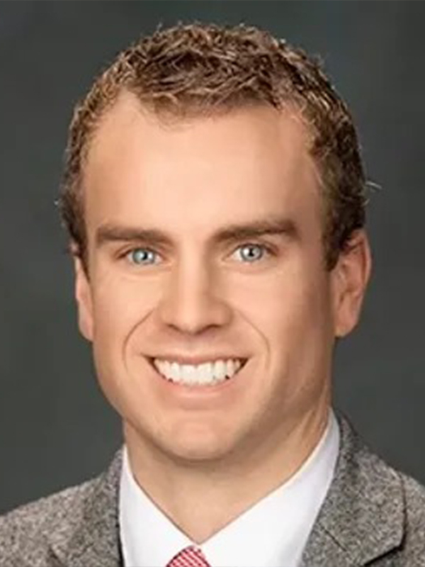 Smiling man with short curly hair in gray suit and red tie, professional headshot on dark background.