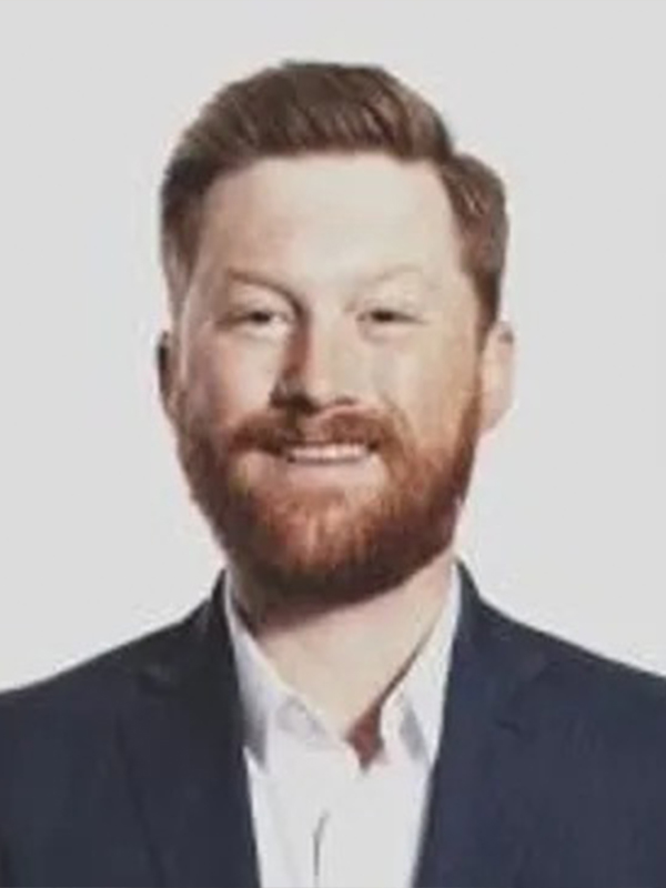 Professional headshot of a smiling man with red hair and beard in a navy blazer and white shirt.