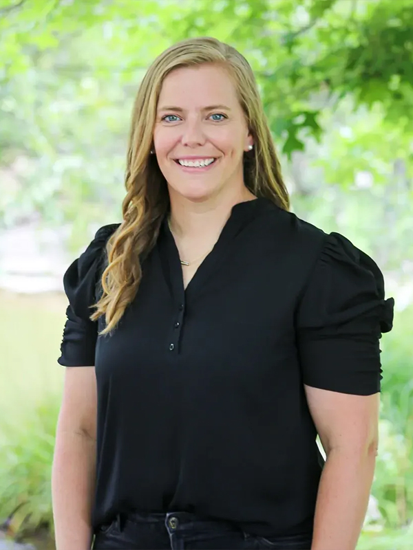 A woman in a black blouse smiling outdoors with leafy green trees in the background.