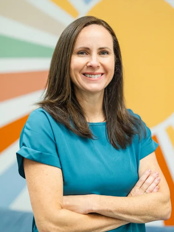 Smiling woman with arms crossed in a teal blouse, standing before a colorful sunburst mural