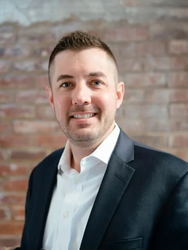 Smiling man in a dark suit and white shirt against a brick wall, professional headshot