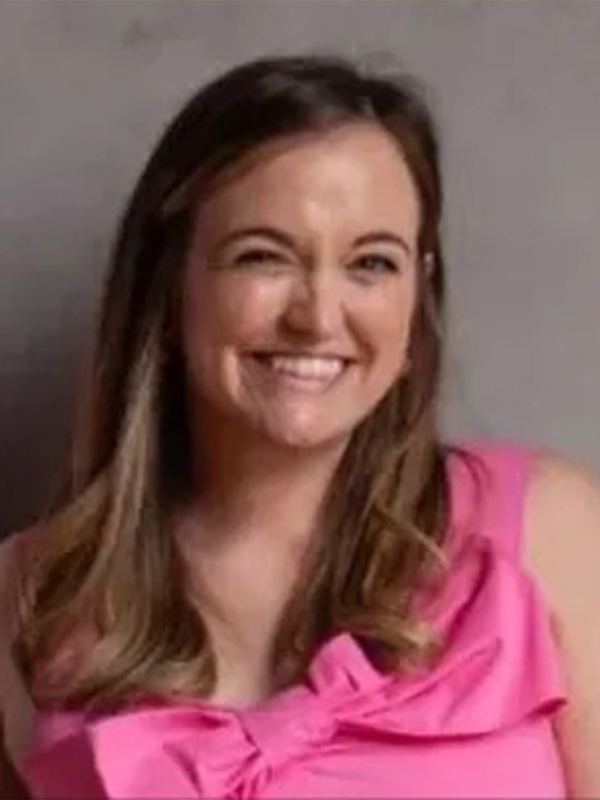 Smiling woman winking in a pink top with a bow, shoulder-length brown hair, posing against a neutral gray background