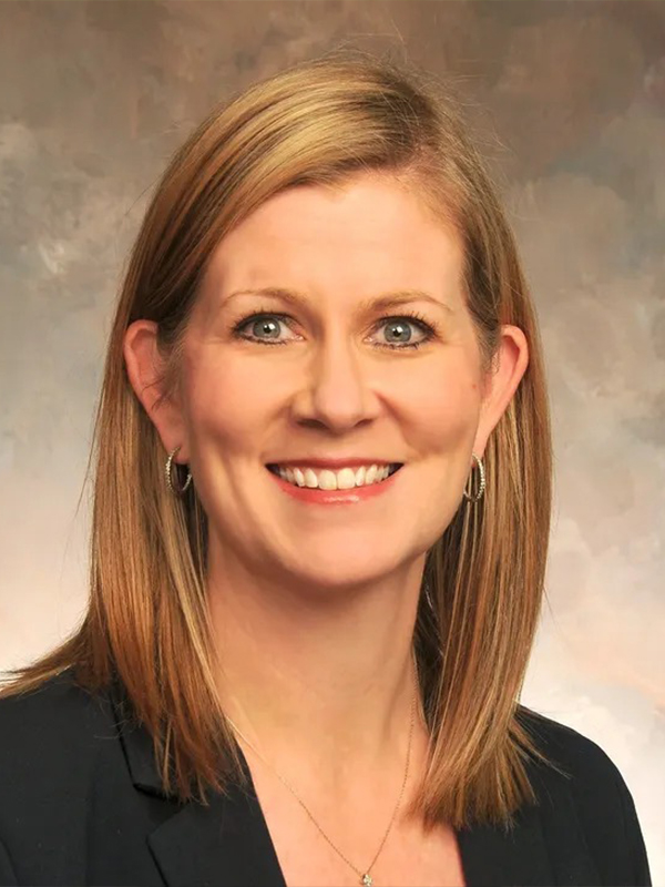 Smiling professional woman in blazer with shoulder-length blonde hair, studio headshot on neutral background.