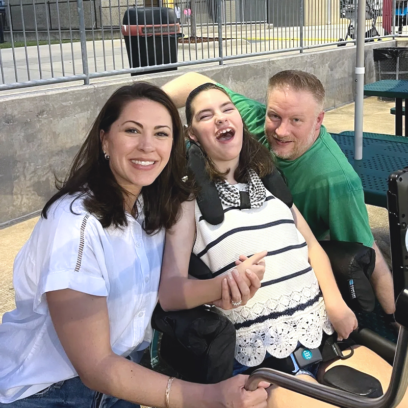 Smiling family with child in a wheelchair at outdoor picnic tables.