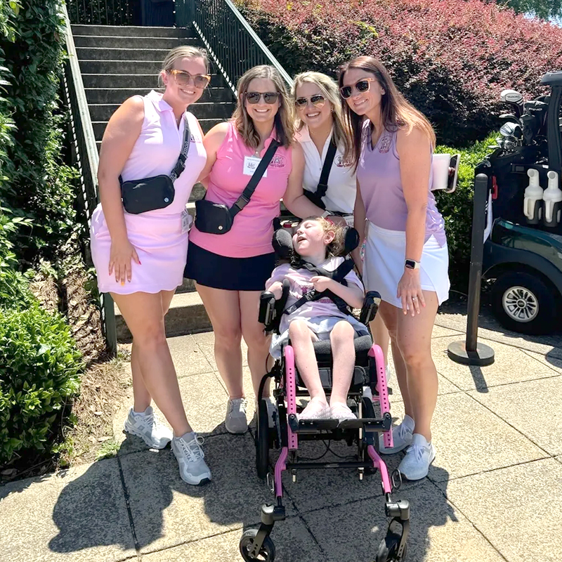 Group of women in golf outfits posing outdoors with a child in a wheelchair beside a golf cart and steps