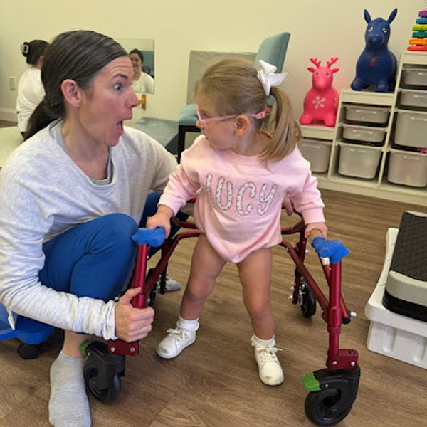 A child using a red walker with a woman therapist assisting in a pediatric therapy room with toys and storage bins.