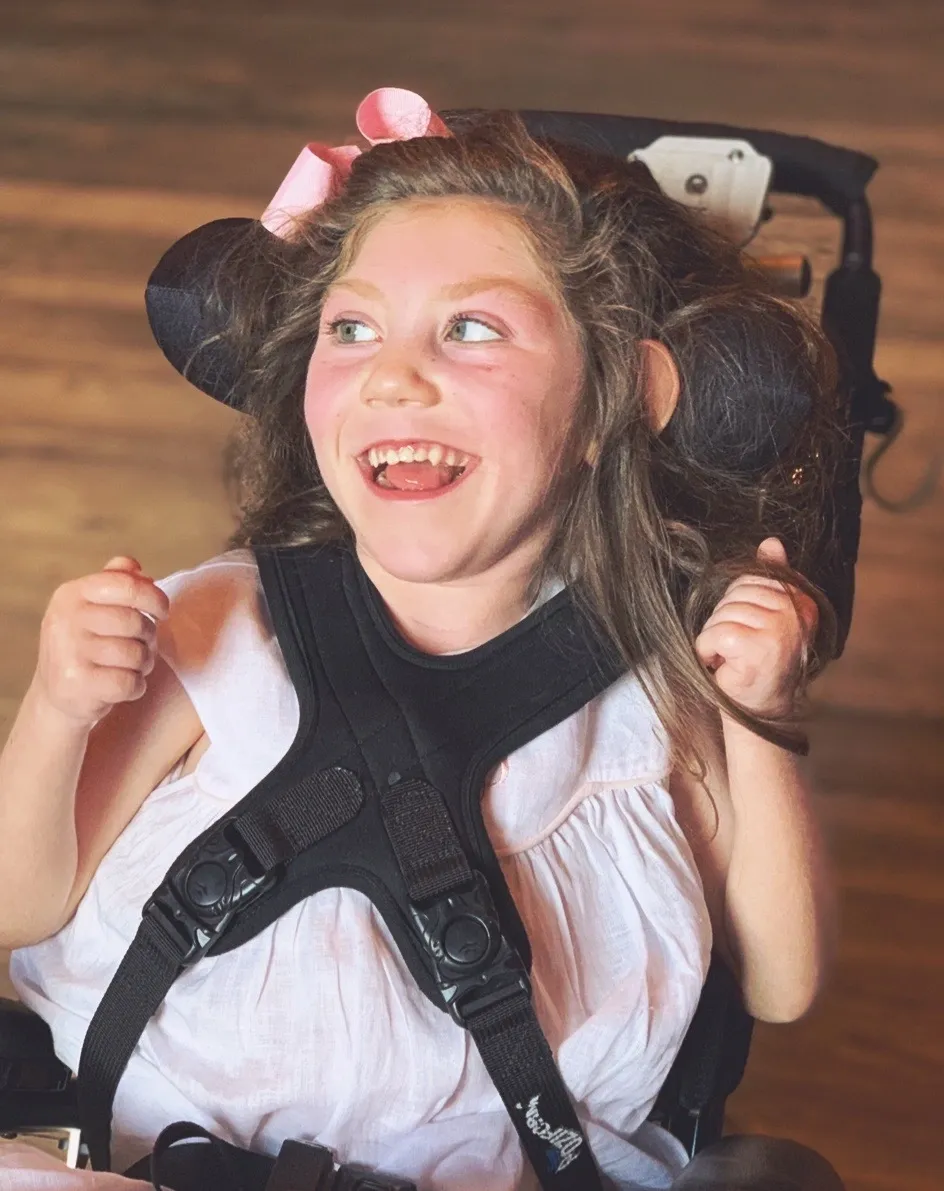 Smiling child with a pink hair bow in a white dress looking to the side indoors, joyful expression