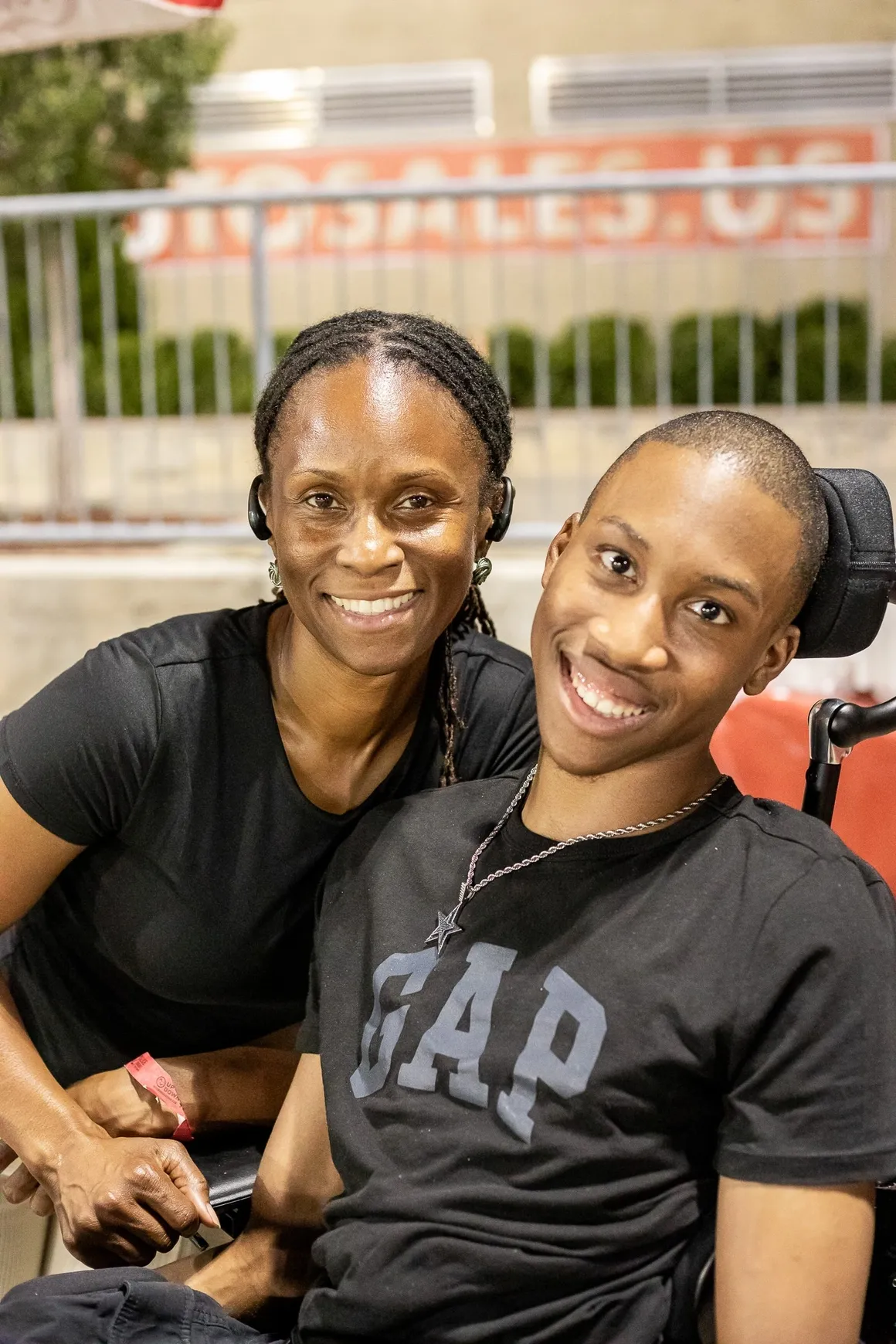 Two people smiling outdoors, one seated in a wheelchair, both wearing black shirts