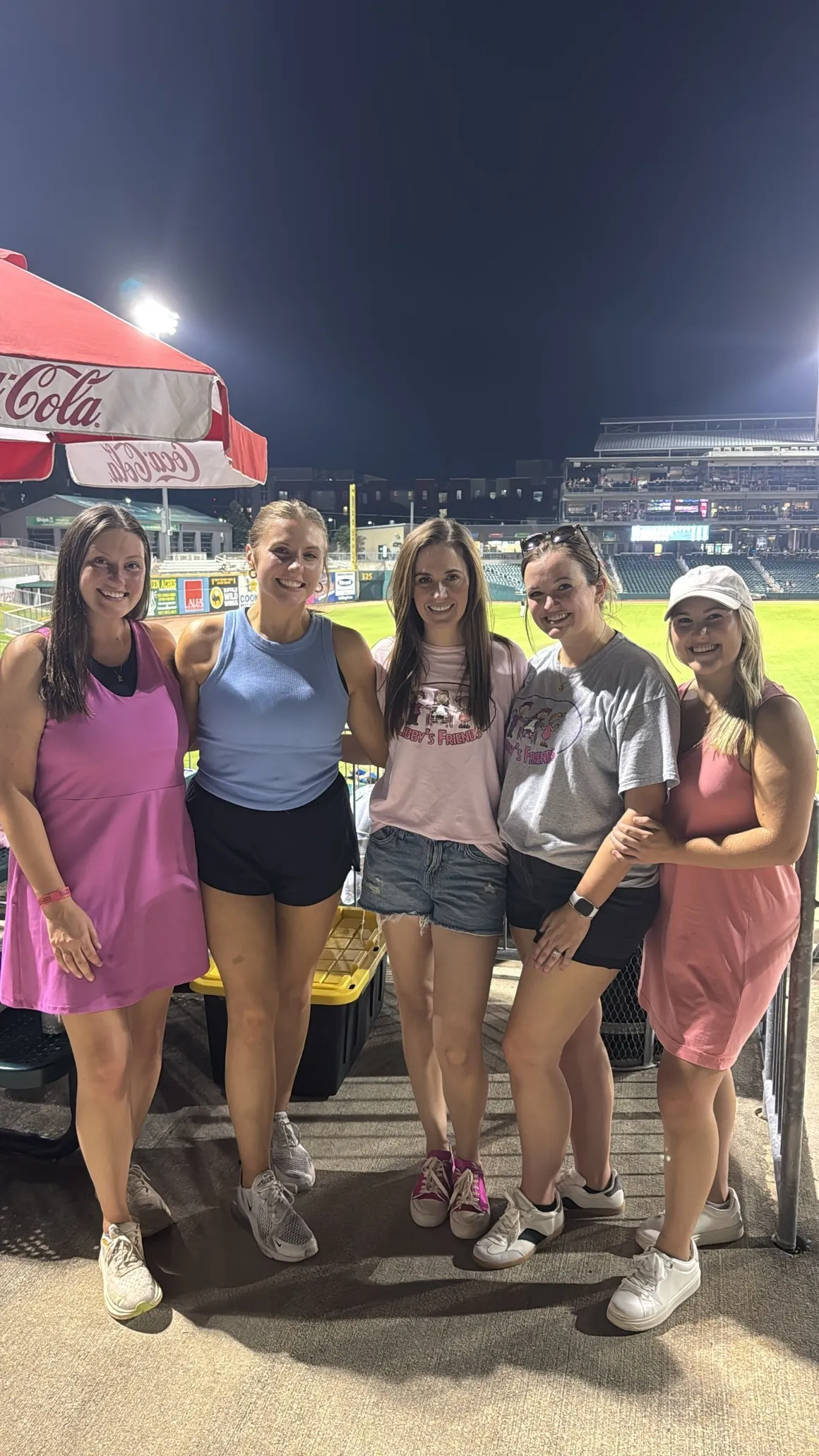 Five women posing at a nighttime baseball stadium with field lights, stands, and a Coca-Cola umbrella in background.
