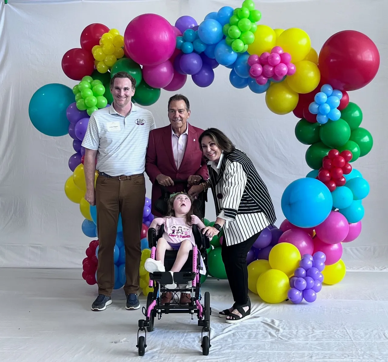 Three adults with a child in a wheelchair posing under a colorful balloon arch on a white backdrop