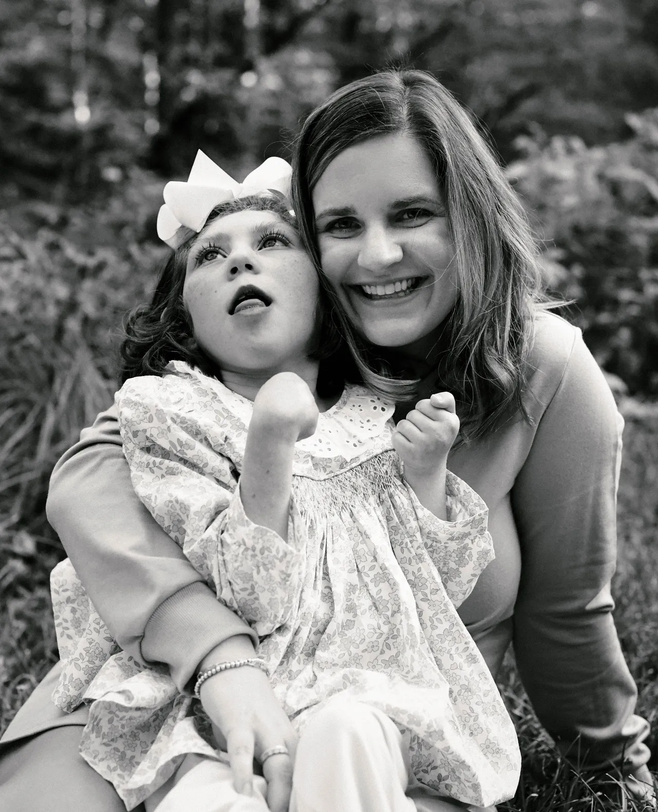 Smiling woman holding a child with a bow in a floral dress, black-and-white outdoor portrait.