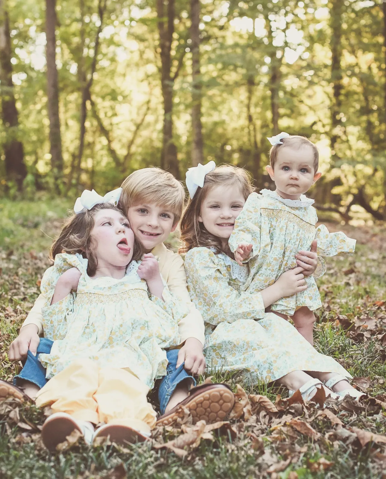 Four children sitting on autumn leaves in a forest, smiling, wearing matching floral outfits and bows.