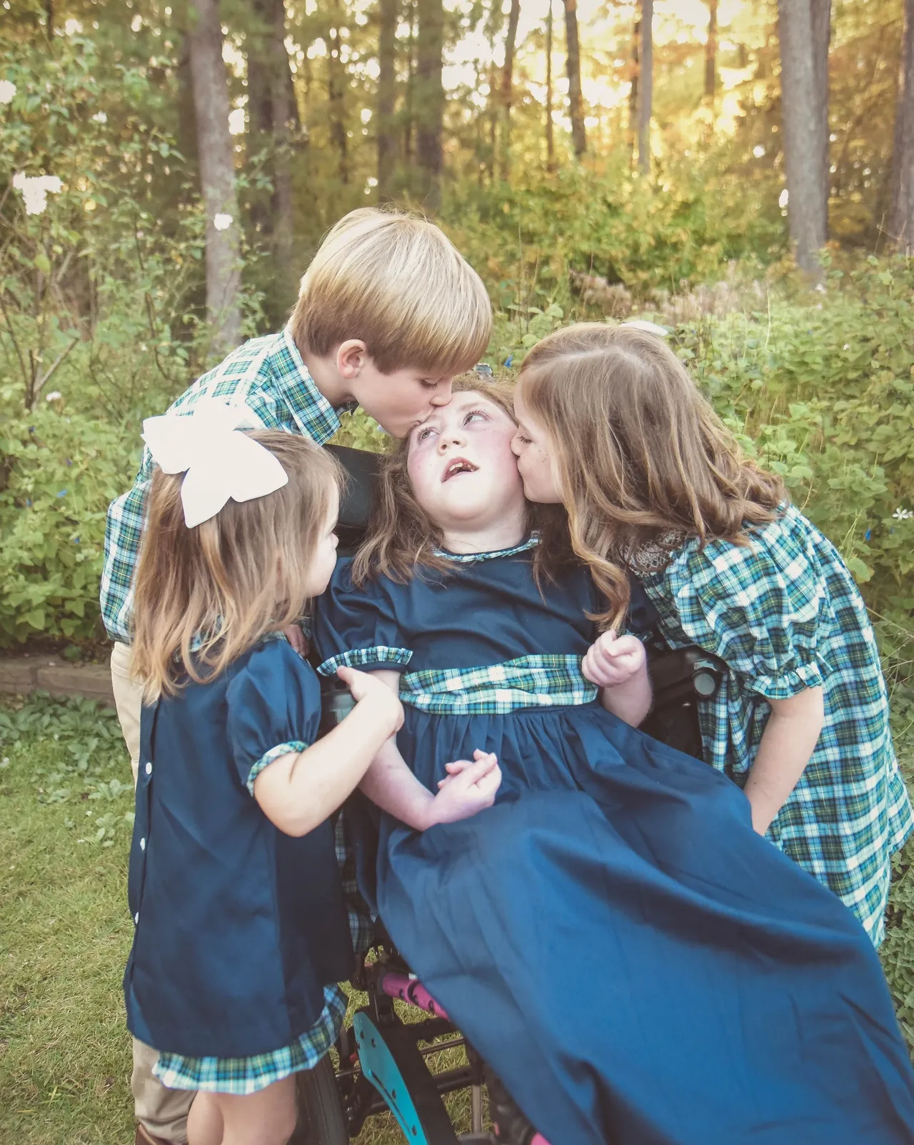Three children kiss a child in a wheelchair during a portrait in a forest garden at sunset, wearing plaid.