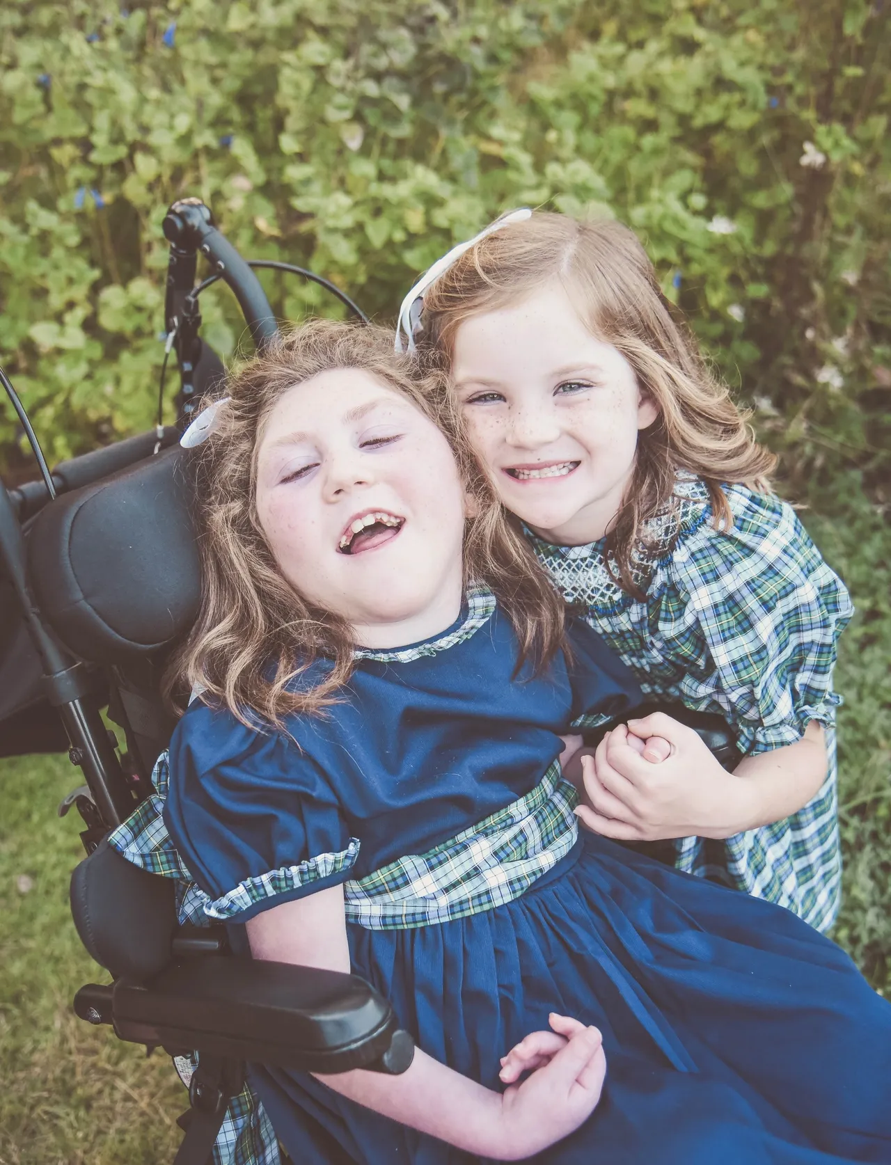 Two girls in blue plaid dresses smiling and hugging in a garden, summer outdoor portrait