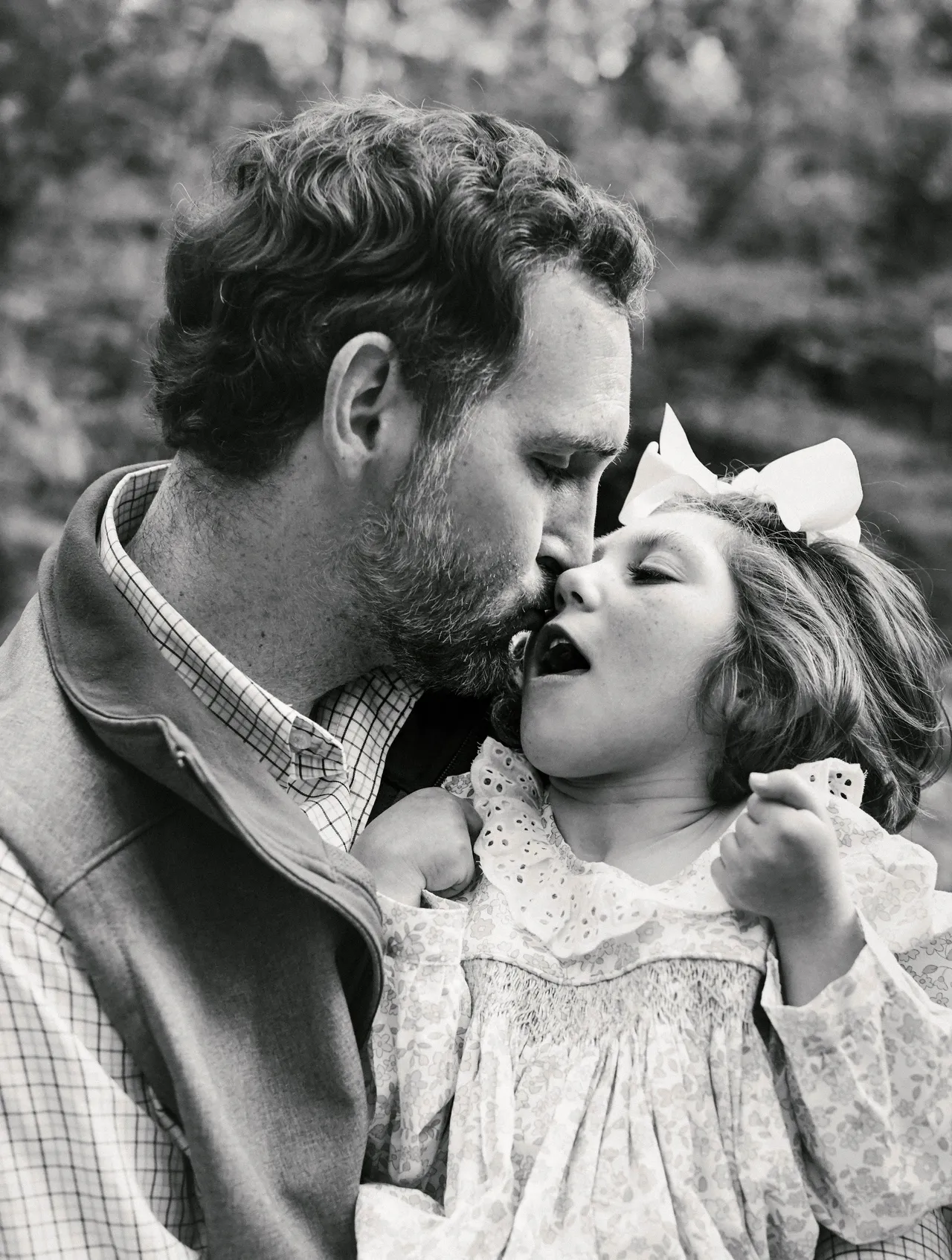 Black-and-white portrait of a man kissing a child with a hair bow outdoors