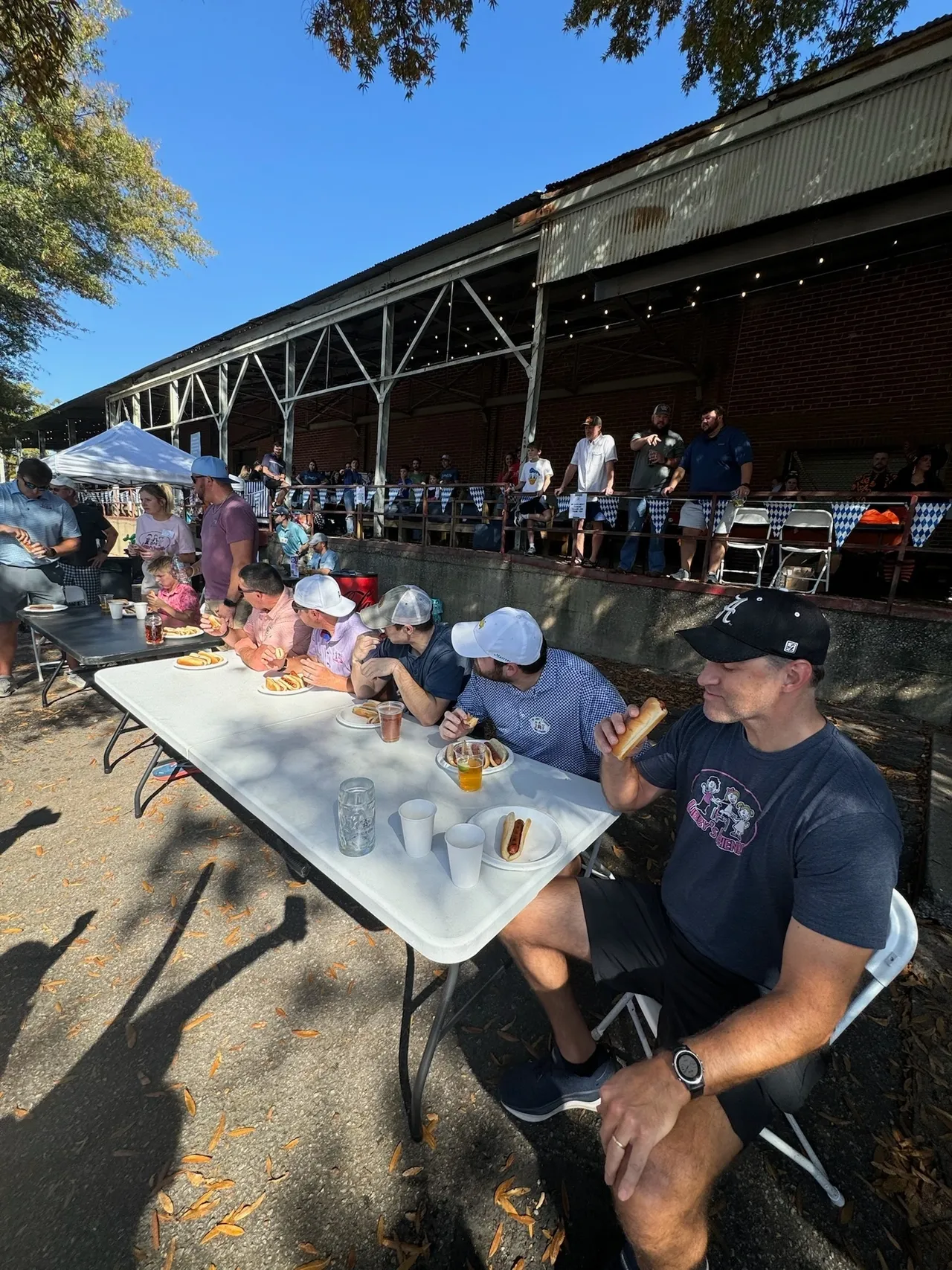 People in a hot dog eating contest at an outdoor festival, seated at a table with spectators by a pavilion.