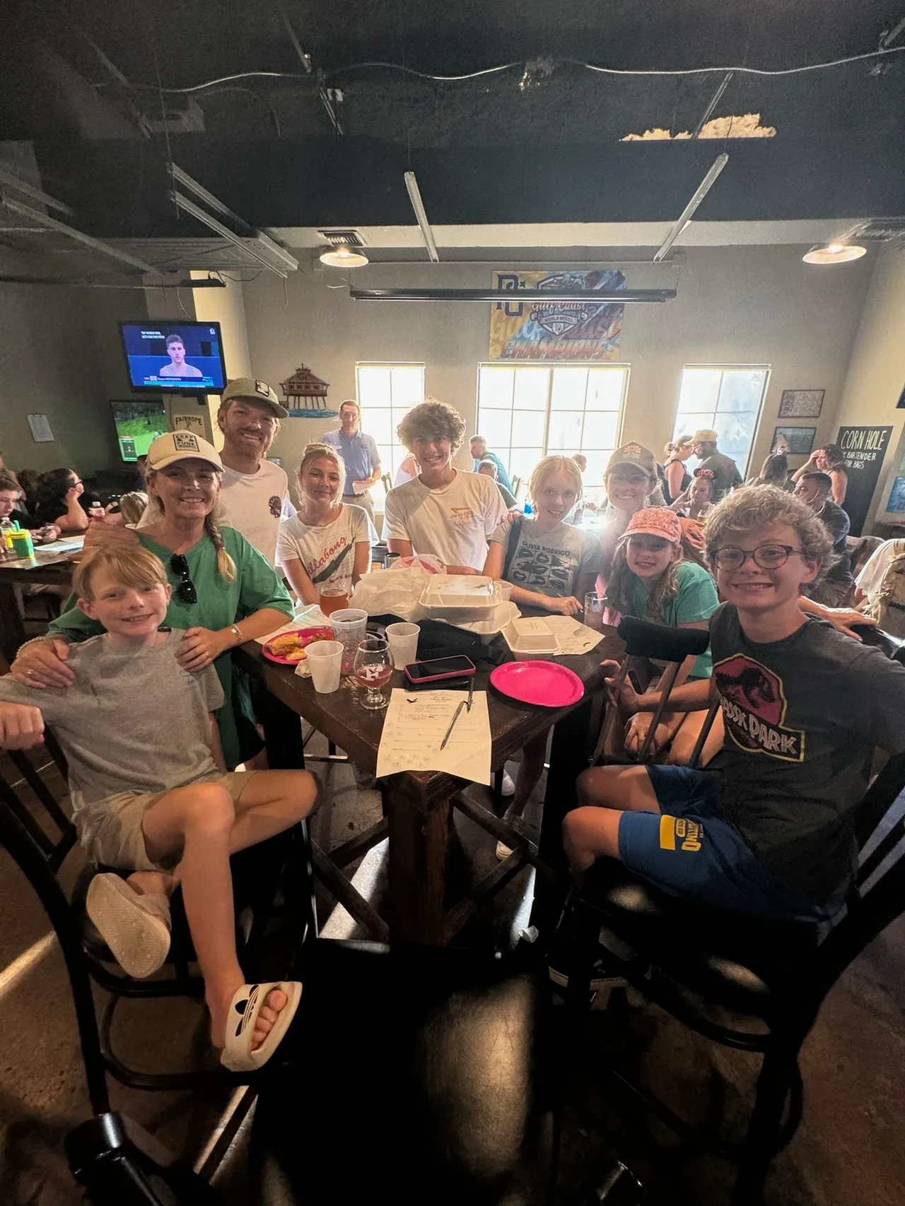 Group of people and kids smiling around a restaurant table during a casual meal in a bright indoor dining space.