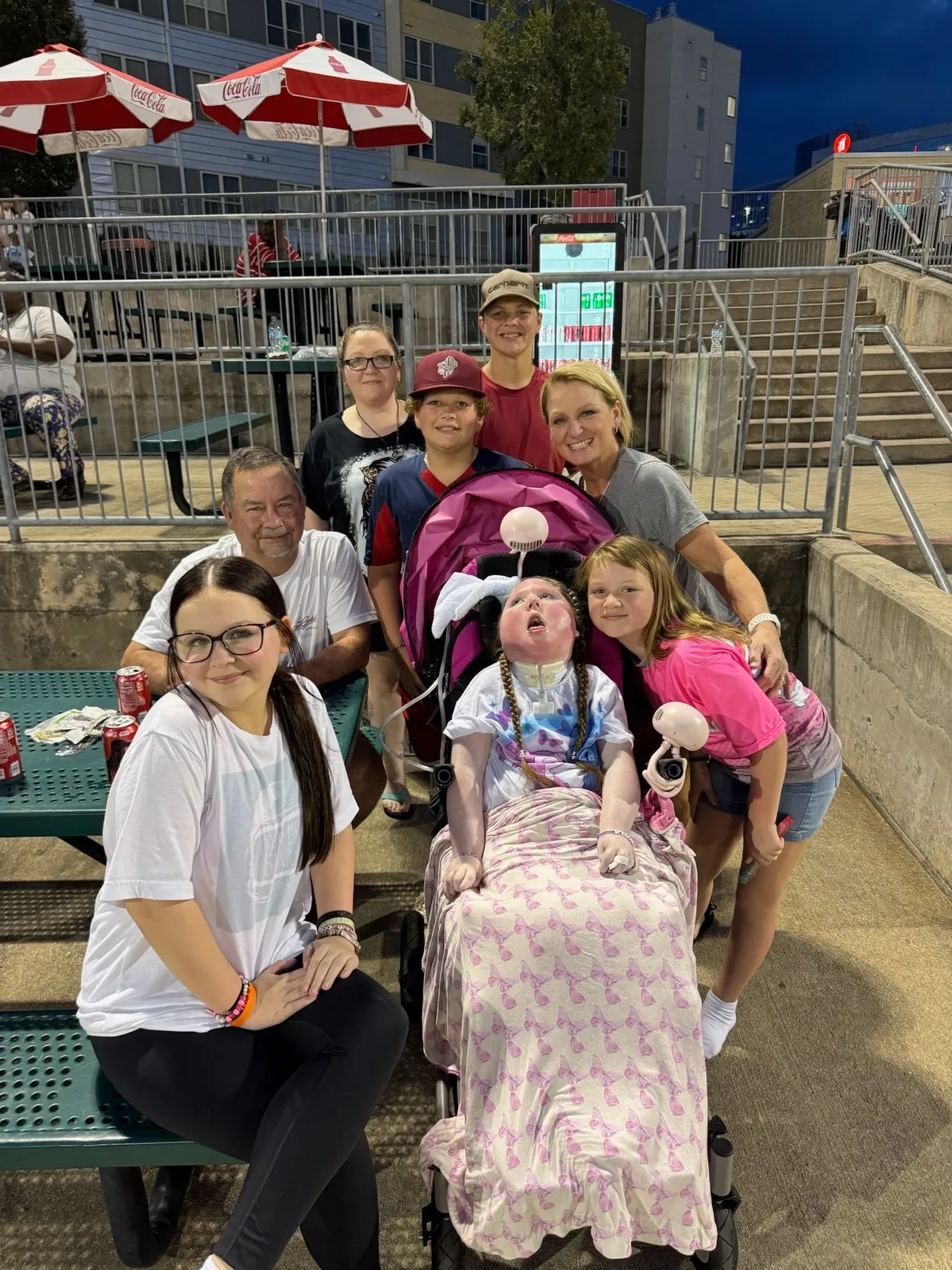 Family group at a ballpark at night, gathered around a person seated in a pink chair with a blanket