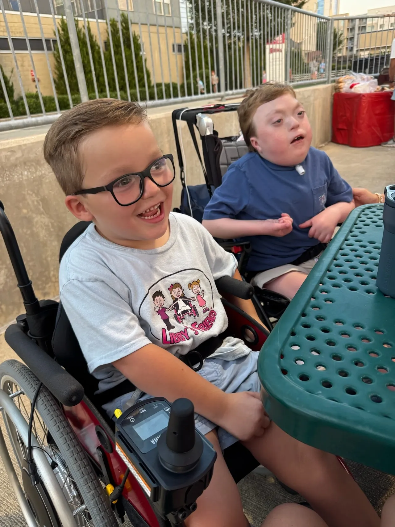 Two children smiling and laughing at an outdoor table with a teal perforated top, urban backdrop and metal railing