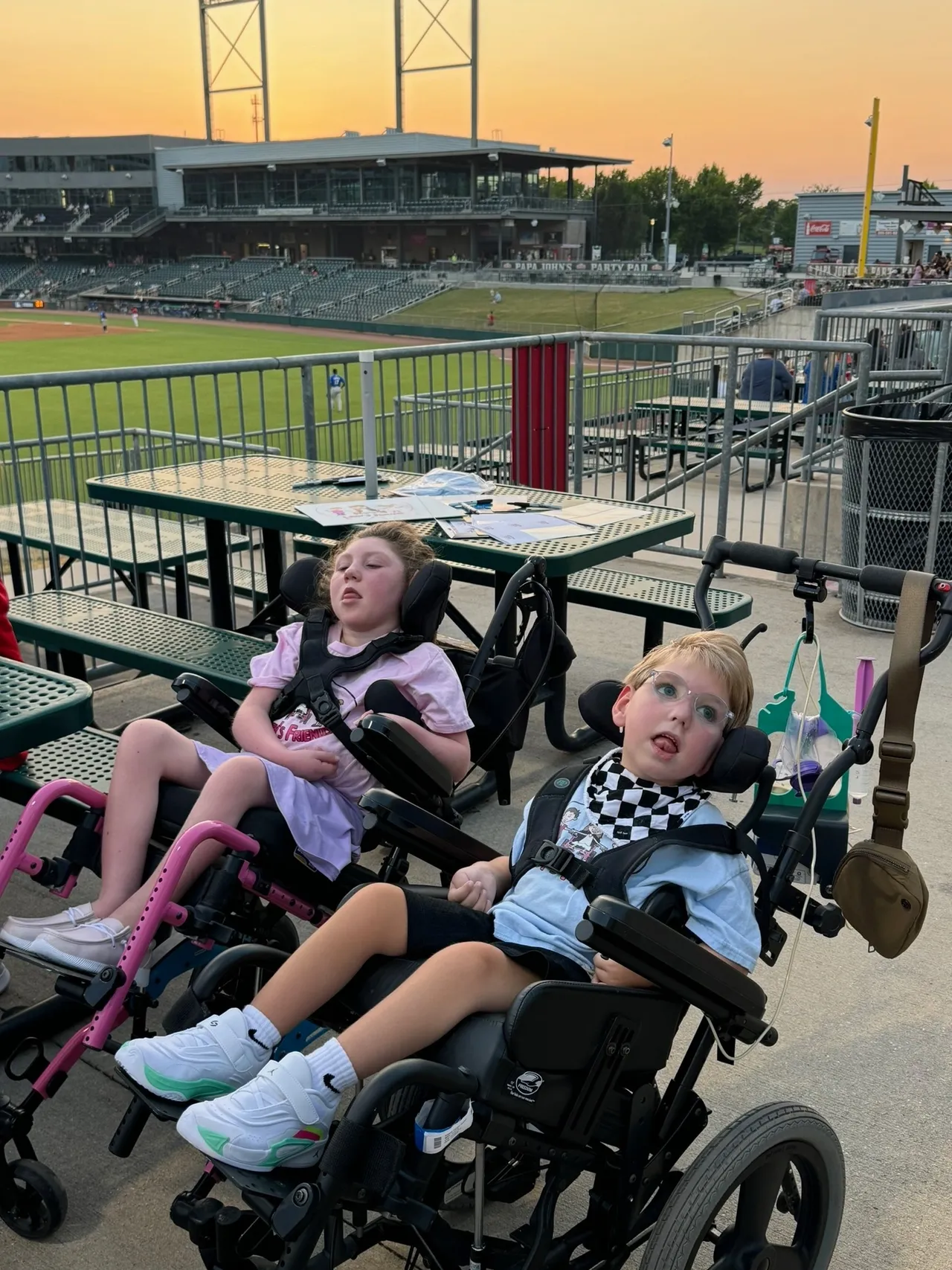 Two children in wheelchairs watching a baseball game from the stands at sunset, with empty seats and field in the background
