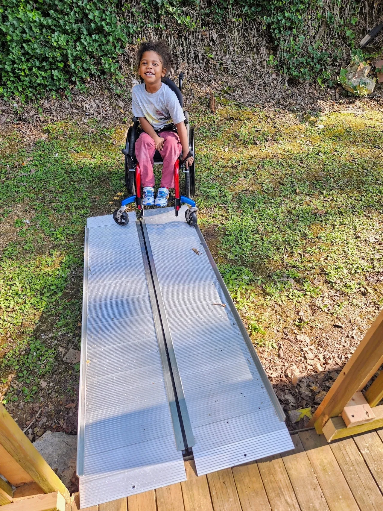 Smiling child in a wheelchair at a portable wheelchair ramp leading to a wooden deck in a sunny backyard.