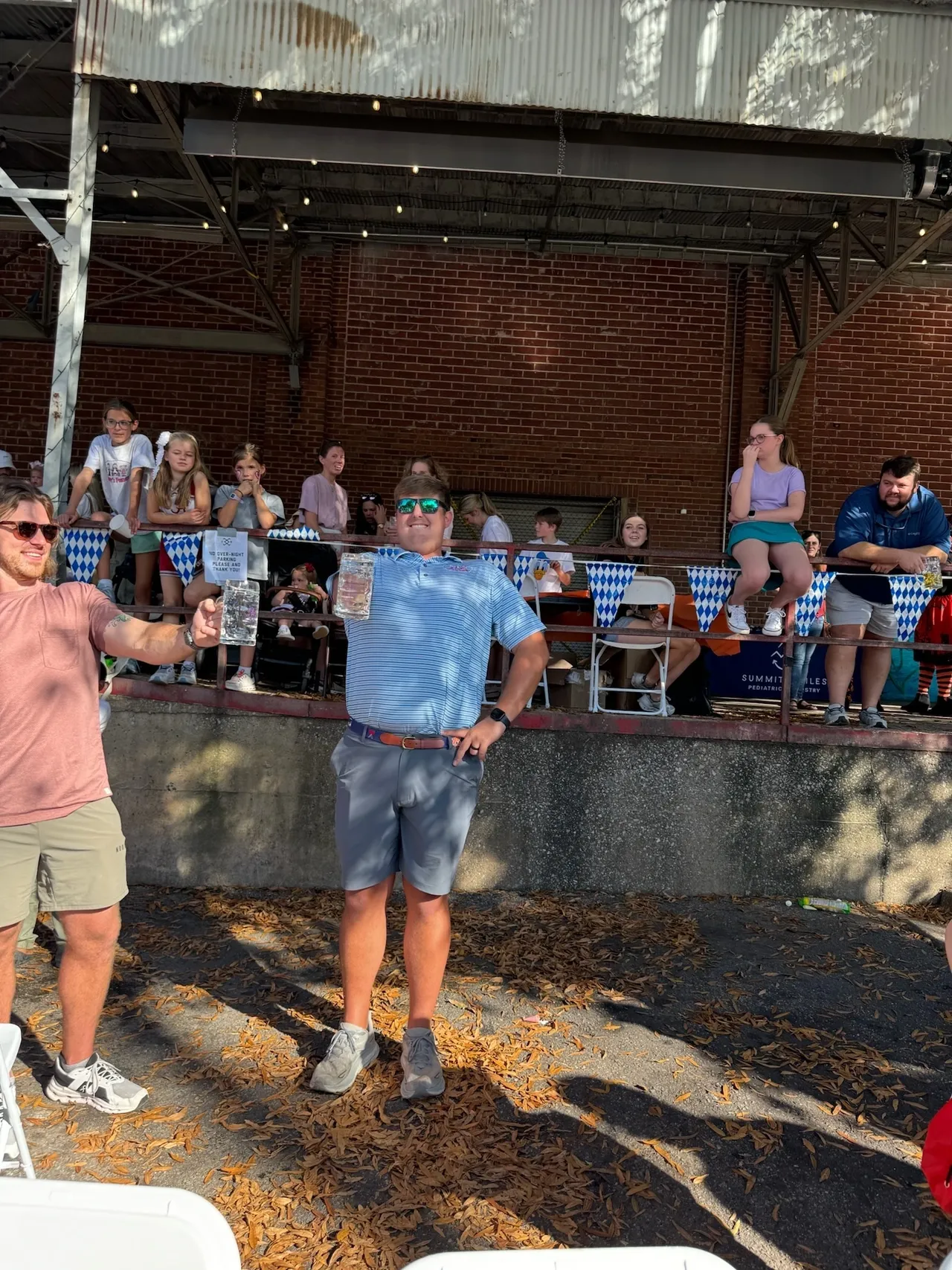 A man in sunglasses holds a beer stein at an outdoor Oktoberfest-style event, with a crowd and blue-check bunting.