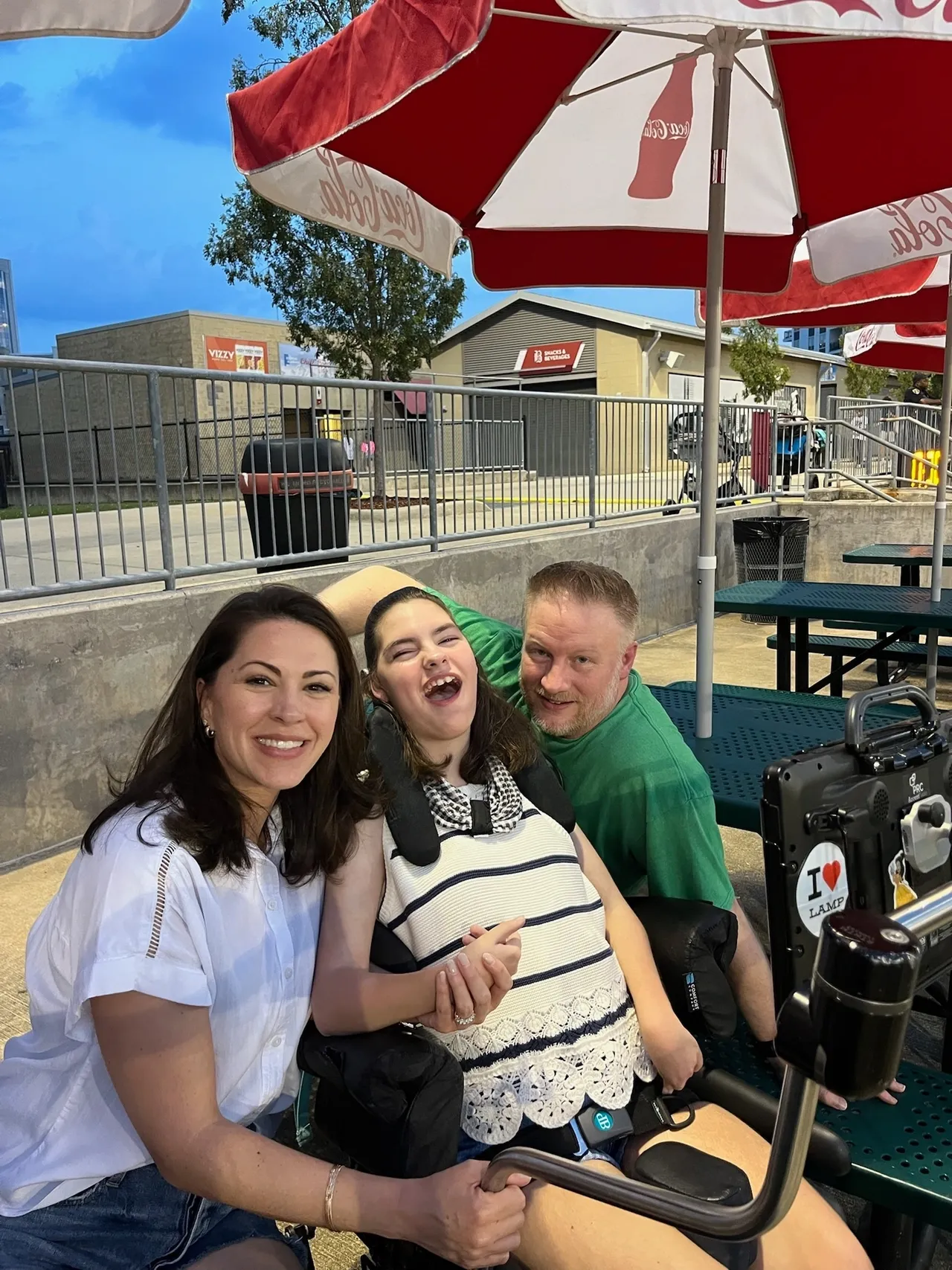 Smiling woman, a person in a wheelchair, and a man at outdoor tables under red Coca‑Cola umbrellas at dusk