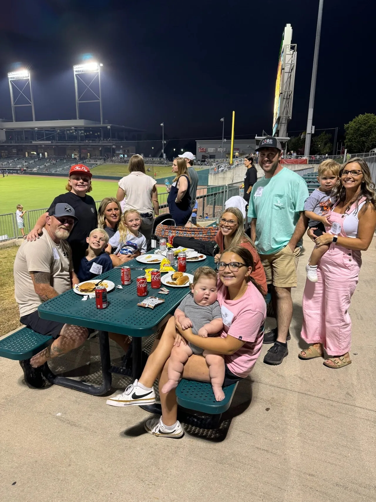 Family group eating at picnic table during night baseball game at stadium, smiling with kids and baby, lights, field behind.