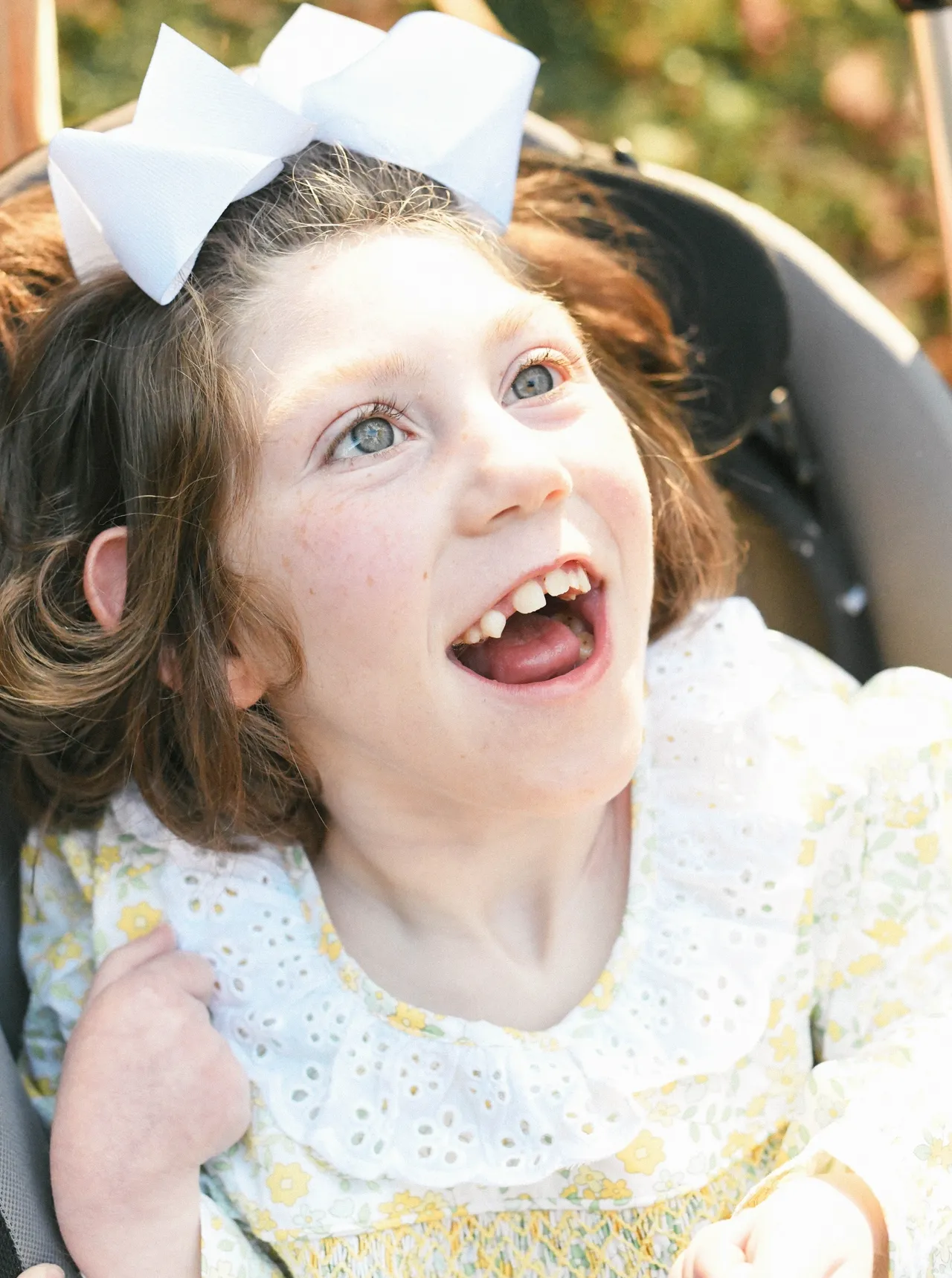 Smiling child with white bow and floral dress looking up while seated outdoors in sunlight